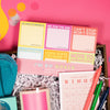 On a hot pink and yellow background, a handful of snarky, colorful, work-themed gifts are arranged in a tray among white paper packing crinkle. Round tissue rainbow confetti is scattered around the tray. This photo is a close-up of the 6-pack of "I'm So Goals" sticky notes and Horrible Meeting Bingo pad. The sticky notes includes six different designs including yellow notes that say "Oh Yeah I'm #Goals," pink notes that say "Me for the Win," and Red notes that say "Can't Stop Won't Stop."
