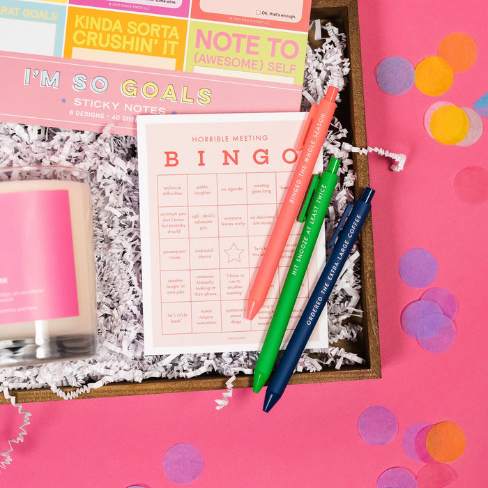 On a hot pink and yellow background, a handful of snarky, colorful, work-themed gifts are arranged in a tray among white paper packing crinkle. Round tissue rainbow confetti is scattered around the tray. This photo is a close-up of Horrible Meeting Bingo pad and a set of three colorful pens" A coral one that says "Binged the Whole Season," a green one that says "Hit Snooze at Least Twice," and a navy one that says "Ordered the Extra Large Coffee." 