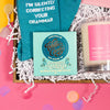 On a hot pink and yellow background, a handful of snarky, colorful, work-themed gifts are arranged in a tray among white paper packing crinkle. Round tissue rainbow confetti is scattered around the tray. This photo is a close-up of the square of Willie's Cacao Sea Kissed Almond Milk Chocolate in a lovely aqua and blue package with gold foil logo.