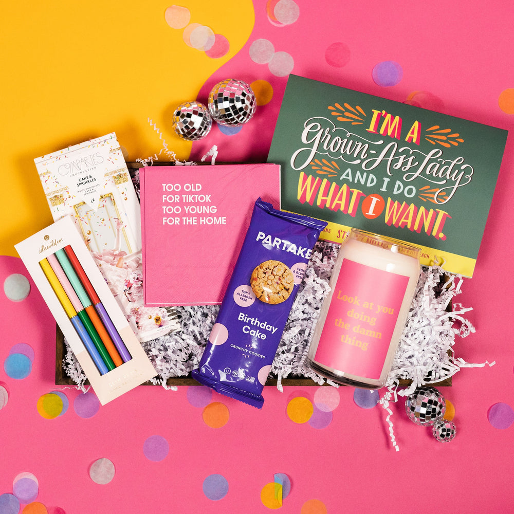 On a hot pink and yellow background, snarky birthday gifts are arranged in a tray with white paper crinkle and confetti. The items include a candle that says "Look at you doing the damn thing", Partake allergy-friendly Birthday Cake crunchy cookies, pink beverage napkins that say "Too Old for TikTok Too Young for the Home," sticky notes that say "I'm a Grown Ass Lady and I Do What I Want," colorful 4-pack of Idlewild pens, and a Compartes cake and sprinkles white chocolate bar.