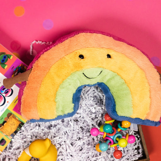 On a hot pink and red background, a handful of colorful baby gifts are arranged in a tray among white paper packing crinkle. Round tissue rainbow confetti is scattered around the tray. The photo is a close-up of the plush rainbow Jellycat with a smiling black simple cartoon face.