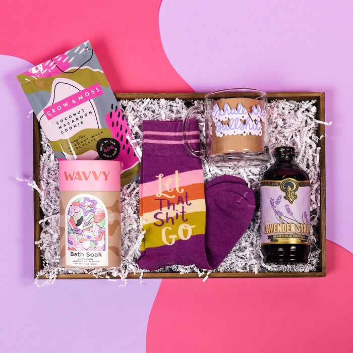 On a hot pink and purple background sits a wooden tray with which paper crinkle and a handful of attractive gifts in a purple color theme. The gifts include a glass mug with lavender script that says "Exhale the Bullshit," purple novelty socks that say "Let that Shit Go," a bottle of Portland Syrups Lavender Syrup premium beverage concentrate, Crow and Moss (dairy free) Coconut Macaroon Cookie and a kraft can of Wavvy Bath Soak in Luscious Lavender and Milk with a cute illustrated label and pink lid.