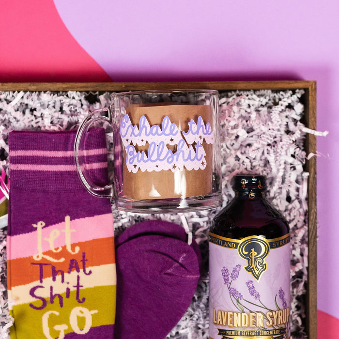 On a hot pink and purple background sits a wooden tray with which paper crinkle and a handful of attractive gifts in a purple color theme. This photo is a close-up of the glass mug with lavender script that says "Exhale the Bullshit," purple novelty socks that say "Let that Shit Go," and 12 oz. bottle of Portland Syrups Lavender Syrup premium beverage concentrate for cocktails or soda water.