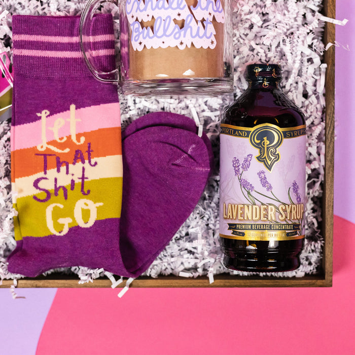 On a hot pink and purple background sits a wooden tray with which paper crinkle and a handful of attractive gifts in a purple color theme. This photo is a close-up of a 12 oz. bottle of Portland Syrups Lavender Syrup premium beverage concentrate for cocktails or soda water, purple novelty socks that say "Let that Shit Go," and a glass mug with lavender script that says "Exhale the Bullshit."