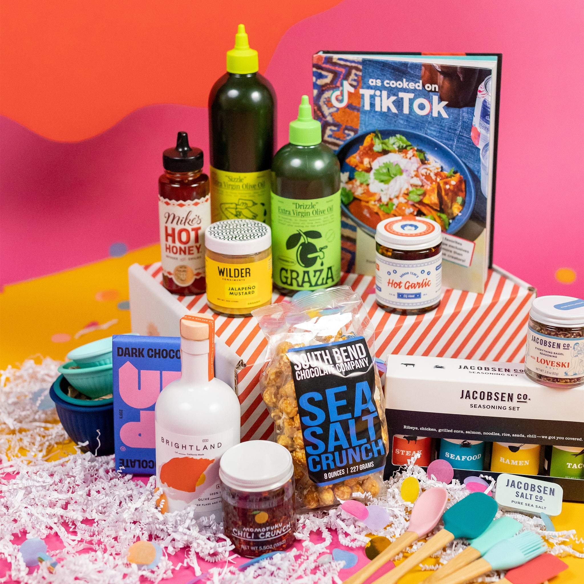 On a pink background with mustard cutout shapes sits a red and white striped box with kitchen gifts stacked around it on white paper crinkle and confetti. The gifts include two Graza olive oils, multicolored silicone utensils, Jacobson Salt Co. flavored salts, Mike's Hot Honey, "As Cooked on TikTok" cookbook, Momofuku Chili Crunch, Silicone finger bowls, Wilder Jalapeño Mustard, Brightland Olive Oil, Mama Teay's Hot Garlic, Up Up Dark Chocolate and South Bend Popcorn Company Sea Salt Crunch Caramel Corn.