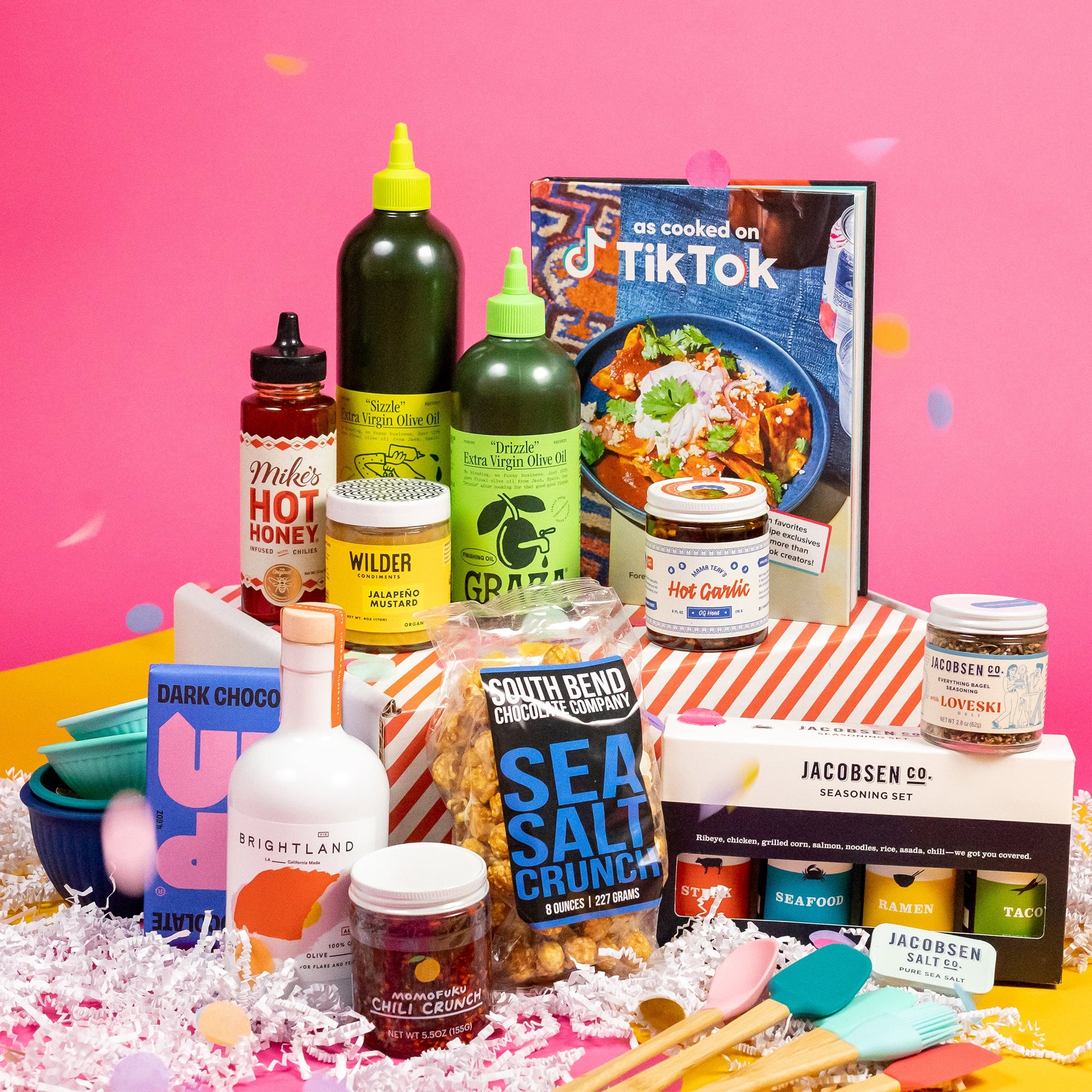 On a pink background with mustard cutout shapes sits a red and white striped box with kitchen gifts stacked around it on white paper crinkle and confetti. The gifts include two Graza olive oils, multicolored silicone utensils, Jacobson Salt Co. flavored salts, Mike's Hot Honey, "As Cooked on TikTok" cookbook, Momofuku Chili Crunch, Silicone finger bowls, Wilder Jalapeño Mustard, Brightland Olive Oil, Mama Teay's Hot Garlic, Up Up Dark Chocolate and South Bend Popcorn Company Sea Salt Crunch Caramel Corn.