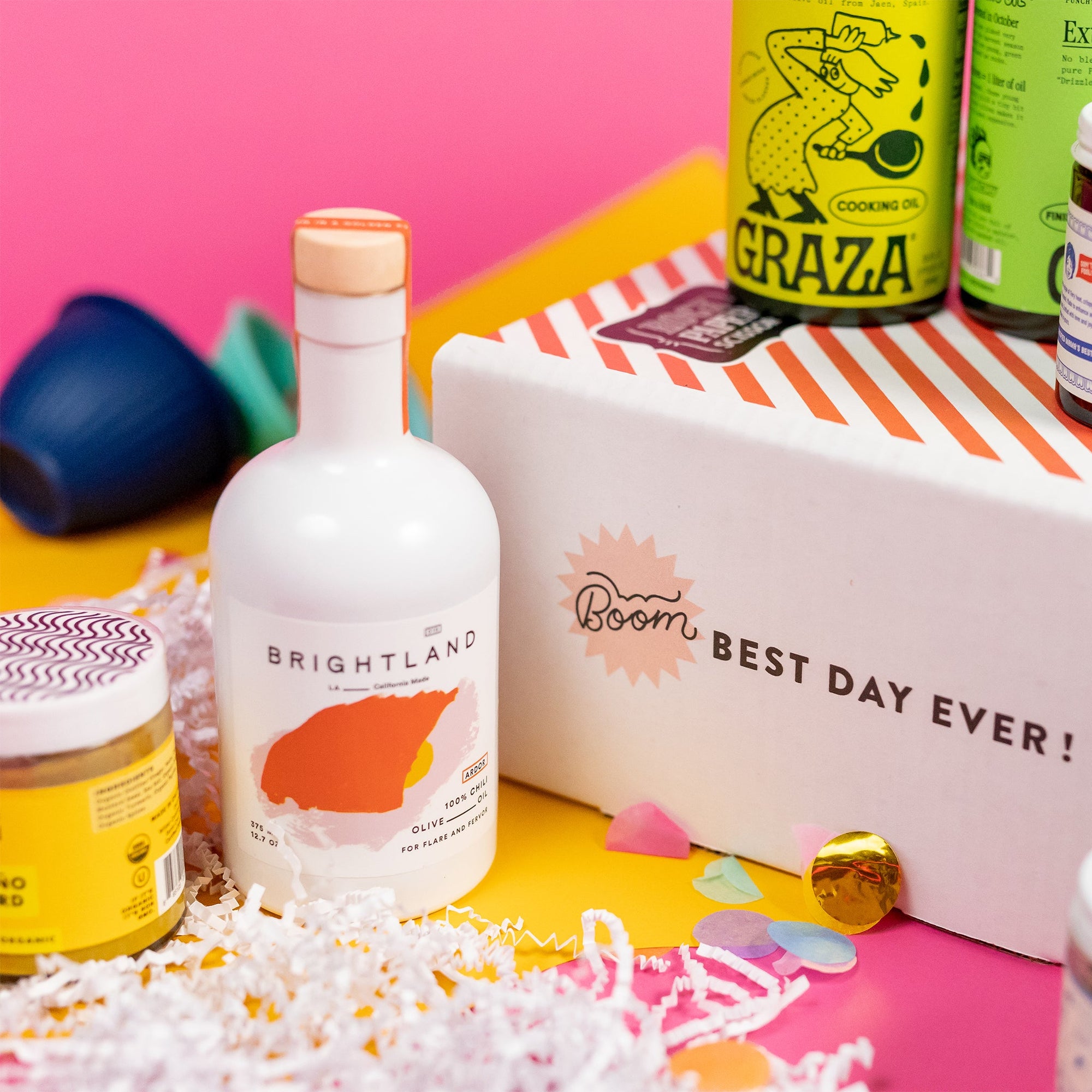On a hot pink background with mustard wobbly cutout shapes sits a red and white striped shipping box with an assortment of lux kitchen accoutrements stacked around it on white paper crinkle with confetti. The side of the box says "BOOM Best Day Ever!" The photo is a close-up of a white bottle of Brightland Olive Oil.