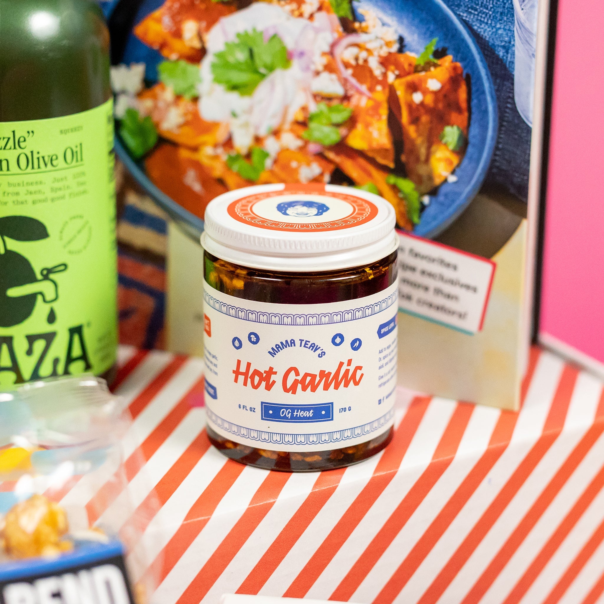 On a red and white striped shipping box sits a jar of Mama Teay's Hot Garlic OG Heat. Behind it, less in focus, is a cookbook and green Graza olive oil.