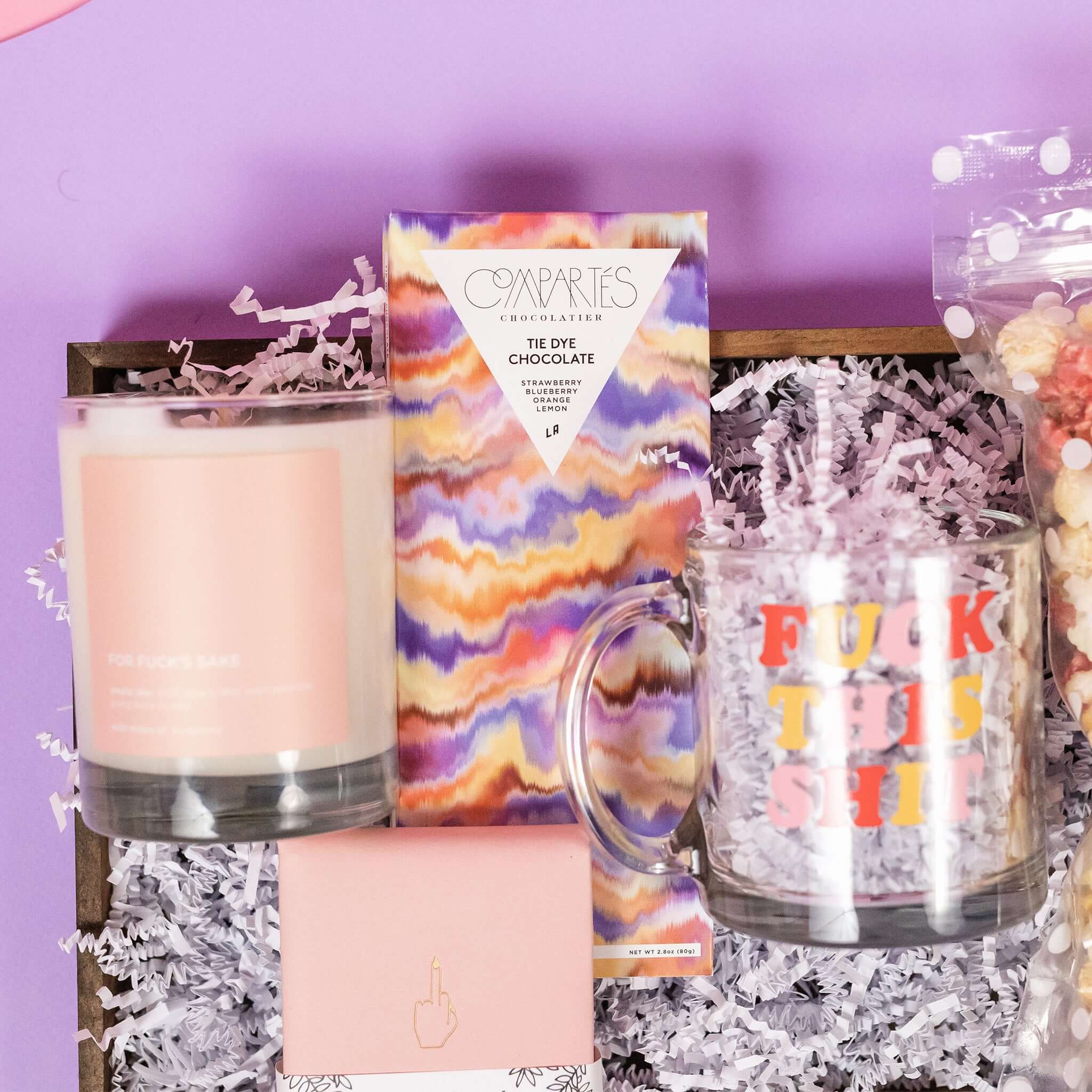 On a baby pink background with lavender wobbly cutout shapes sits a wooden tray with an assortment of pink and lavender 'fuck this shit' themed items and white paper crinkle. This is a close-up on the lux soy candle with baby pink label and white writing "FOR FUCK'S SAKE" and gorgeous Compartes marble chocolate bar.