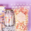On a lavender background sits a wooden tray with an assortment of pink and lavender 'fuck this shit' themed items and white paper crinkle. This photo is a close-up on the glear glass mug that says "FUCK THIS SHIT" in cute alternating red, pink and yellow letters" and clear polka dot bag of Strawberry Shortcake popcorn from the Popcorn Shop.