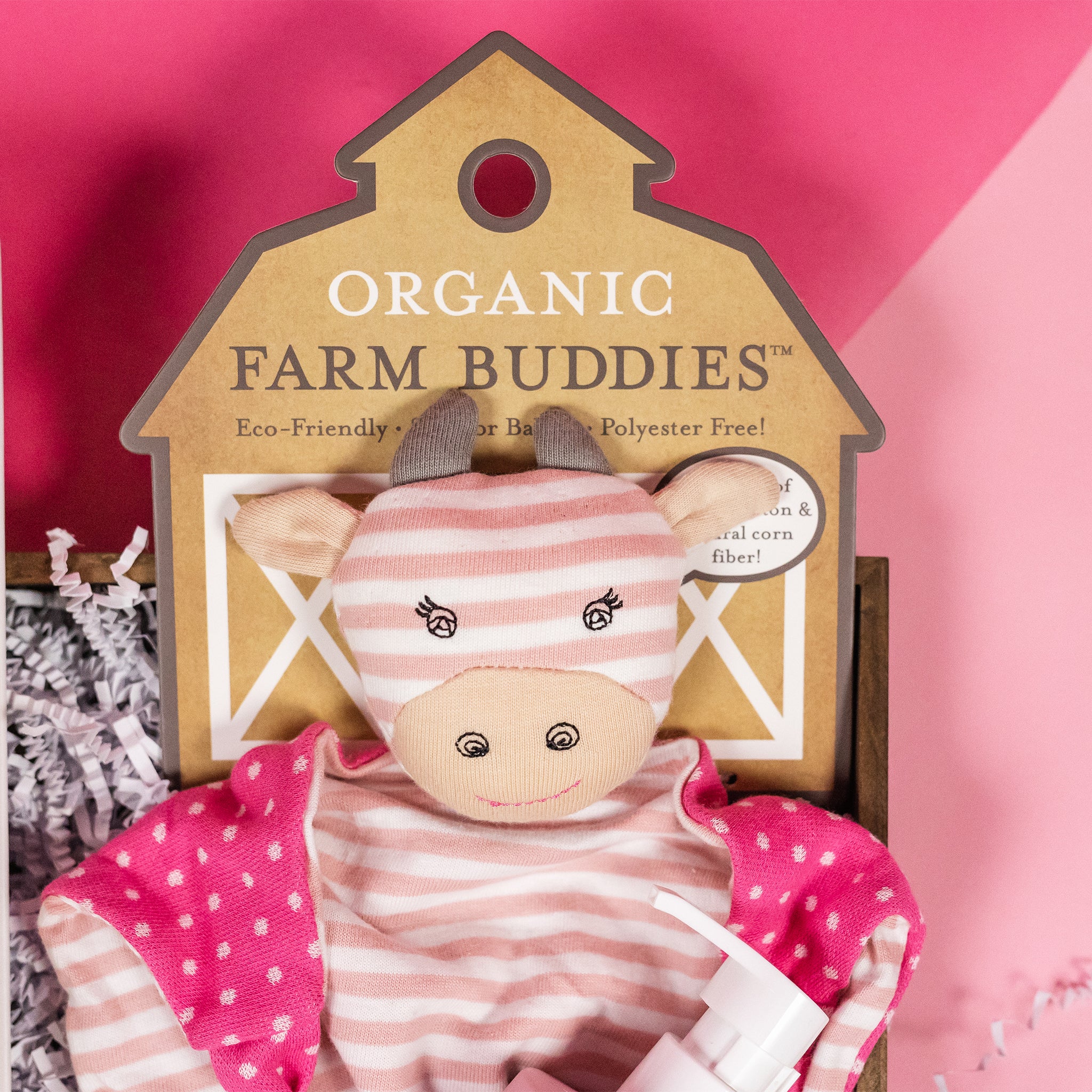 On a background that's split diagonally with hot pink and bubblegum pink sits a wooden tray with white krinkle paper. This is a  close-up of the Organic Farm Buddy pink striped cuddly cow for newborn.