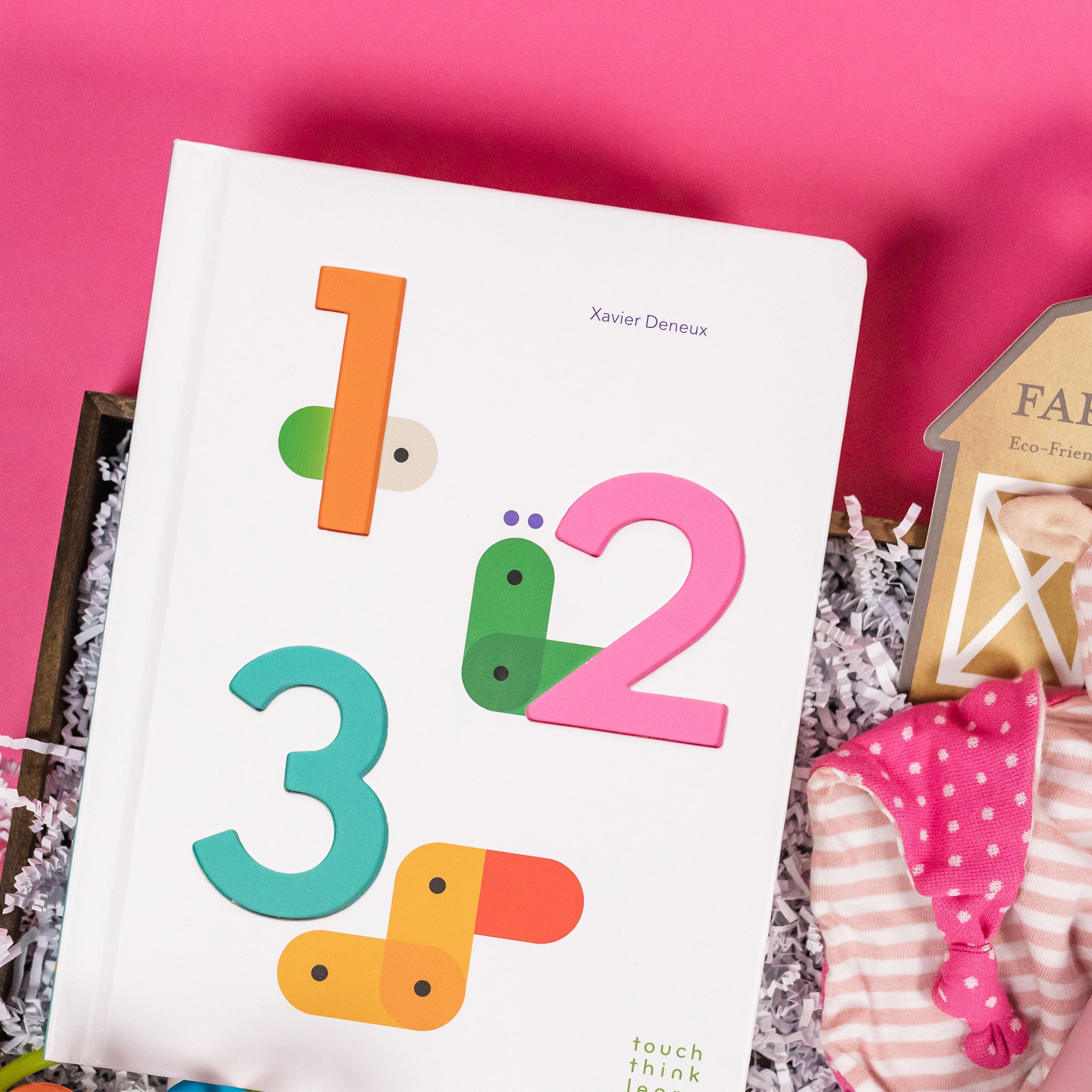 On a hot pink background sits a wooden tray with a few cute pink baby products on white paper krinkle. This is a close-up featuring the 1-2-3 "Touch-Think-Learn" board book by Xavier Deneux.