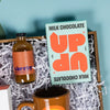 On a calming blue background with green wobbly cutout shape sits a wooden tray of products and white paper krinkle. The products are all in a fun, bright orange theme.  This photo is a close-up on the UP UP 4.6 oz milk chocolate bar.