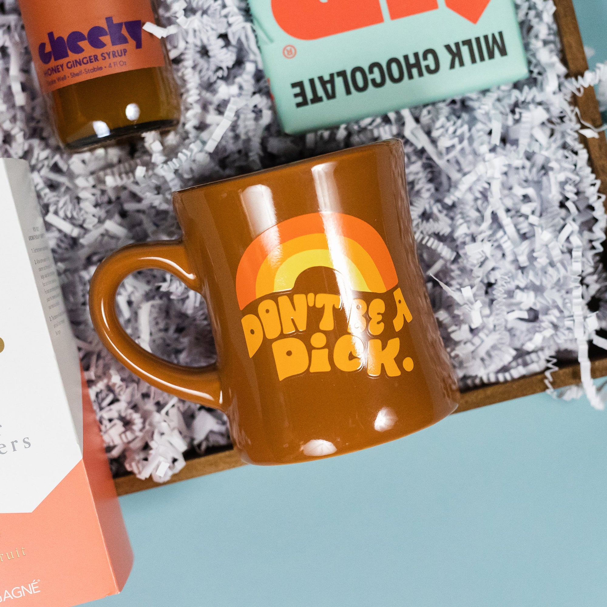 On a calming blue background with green wobbly cutout shape sits a wooden tray of products and white paper krinkle. The products are all in a fun, bright orange theme. This photo is a close-up on the brown diner mug that says "Don't be a Dick" in a retro orange font with a retro orange rainbow above it.