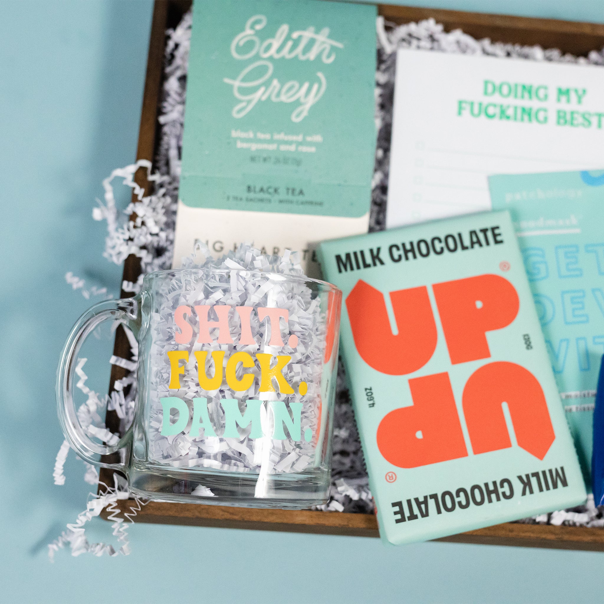 On a calming blue background sits a wooden tray of products and white paper krinkle. The products are a lovely sky blue and aqua theme. This photo is a close-up of the clear glass mug that says "Shit Fuck Damn" in cute pink yellow and aqua words, Edith Grey Black Tea from Big Heart Tea, and UP UP 4.6 oz milk chocolate bar.