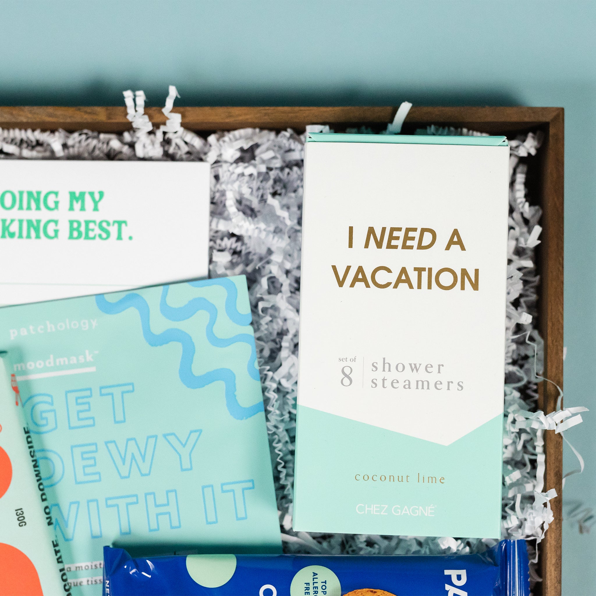 On a calming blue background sits a wooden tray of products and white paper krinkle. The products are a lovely sky blue and aqua theme. This photo is a close-up on the 8 pack of Chez Gagne coconut lime shower steamers that say "I Need a Vacation" and  Patchology moisturizing sheet mask "Get Dewy With It."