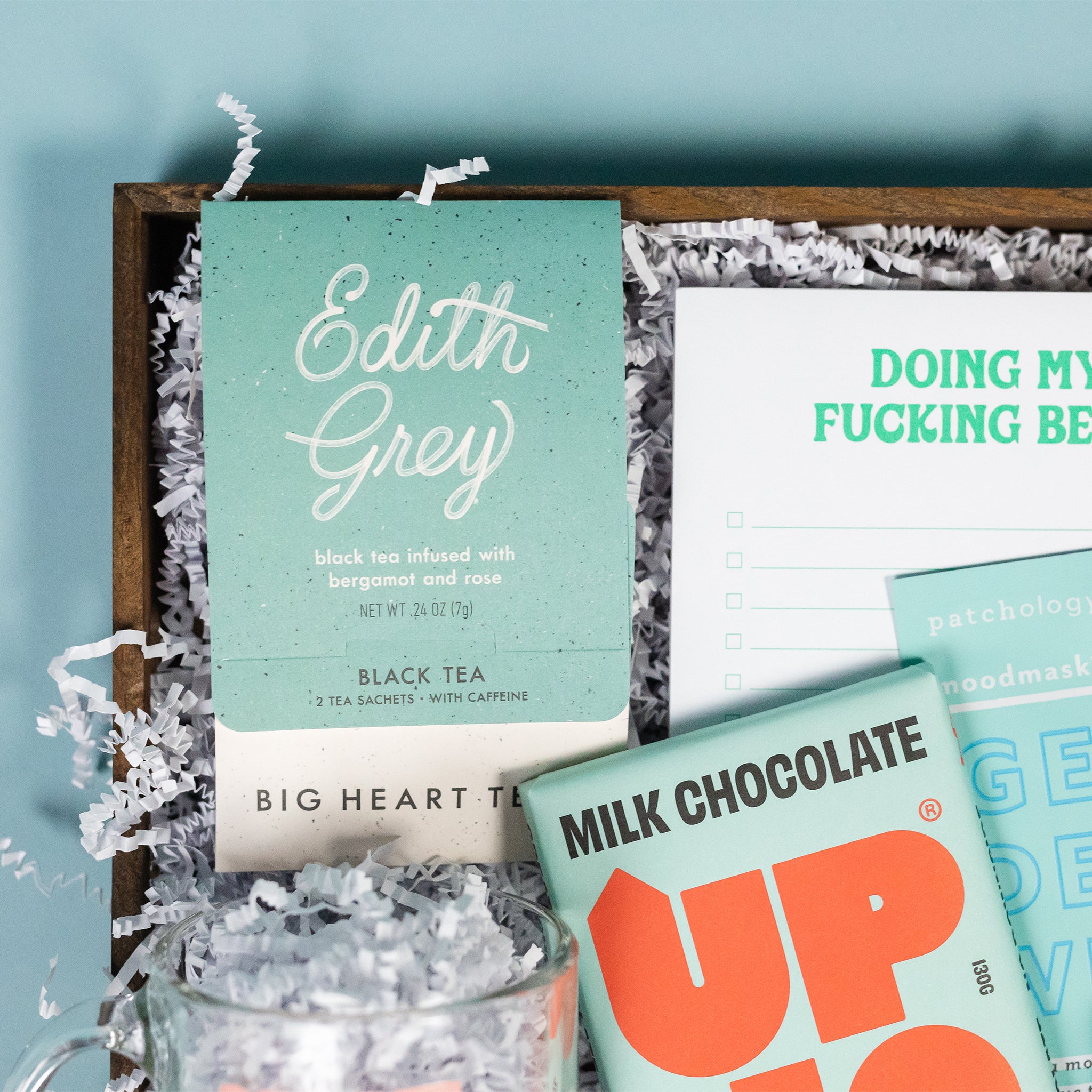 On a calming blue background sits a wooden tray of products and white paper krinkle. The products are a lovely sky blue and aqua theme. This photo is a close-up of the Edith Grey Black Tea from Big Heart Tea in its cute aqua packaging with hand-lettered text.