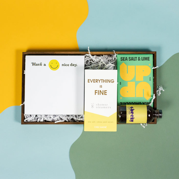 On a calming blue and green background with sunny yellow cutout sits a wooden tray of four products and white paper krinkle. Each product has some sunny yellow on the label. There's a white square notepad with smiley face that says "Have a Nice Day," a pack of 8 Chez Gagne Sea Salt and Neroli "Everything is Fine" shower steamers, UP UP sea salt and lime milk chocolate bar, and mini bottle of Cheeky Honey Syrup.