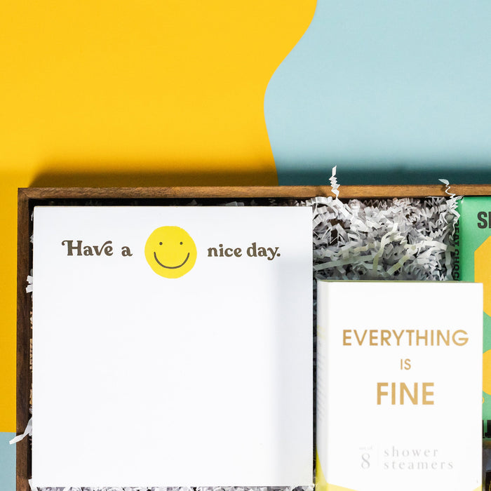 On a calming blue and green background with sunny yellow cutout sits a wooden tray of four products and white paper krinkle. Each product has some sunny yellow on the label. This is a close-up on the white square notepad with smiley face that says "Have a Nice Day." 