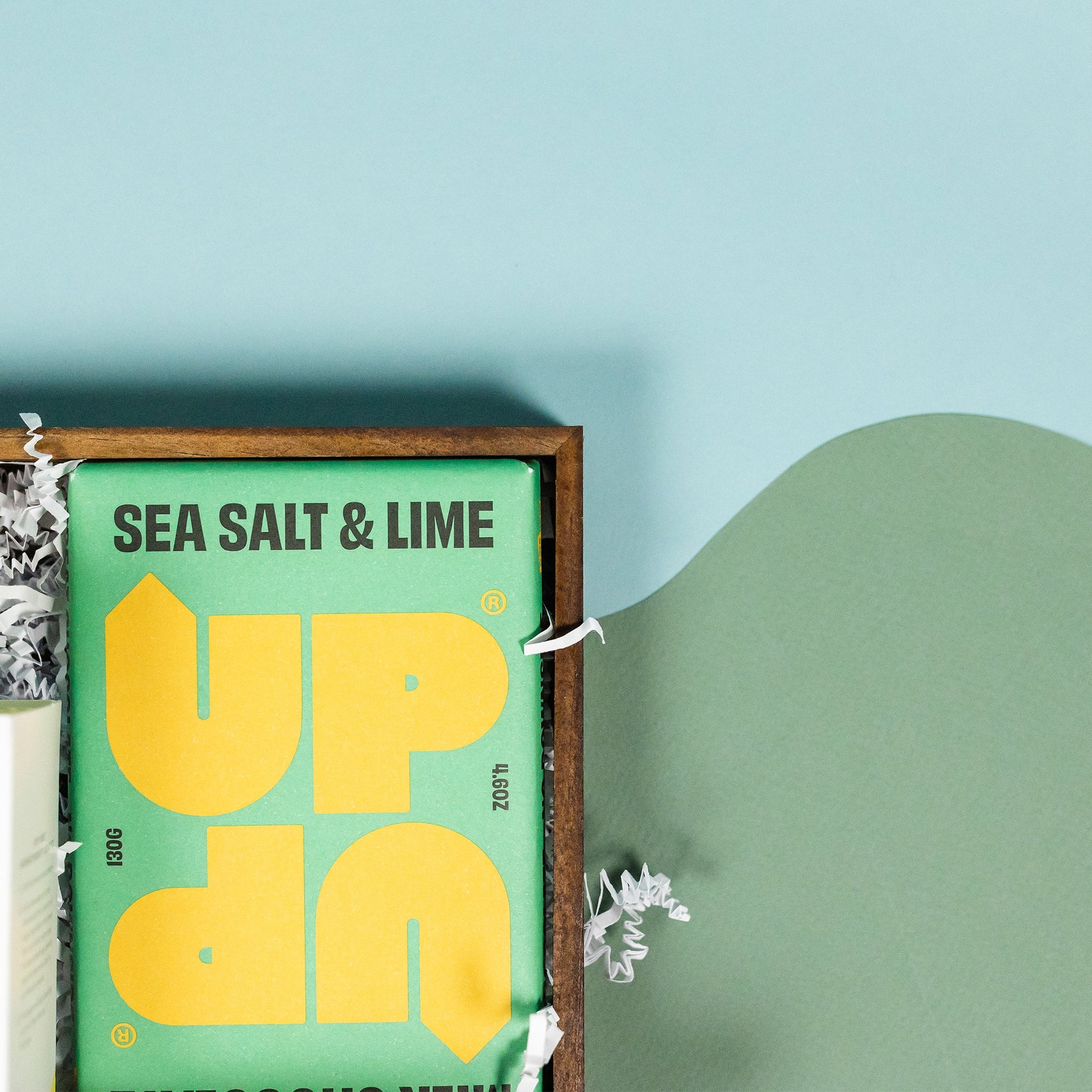 On a calming blue and green background with sunny yellow cutout sits a wooden tray of four products and white paper krinkle. This photo is a close-up on the UP UP sea salt and lime milk chocolate bar. It has a bright grass green label with UP UP in prominent yellow letters.