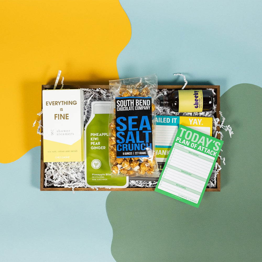 On a calming blue and green background with sunny yellow cutout sits a wooden tray of products and white paper krinkle. Each product has some sunny yellow, green or blue on the label. There's a pack of 8 Chez Gagne Sea Salt and Neroli "Everything is Fine" shower steamers, Pineapple Kiwi Pear Ginger Pineapple Bliss face mask, a mini bottle of Cheeky Honey Syrup, optimistic sticky note set, "Today's Plan of Attack" check box note pad, and a bag of South Bend Chocolate Company Sea Salt Crunch caramel corn.