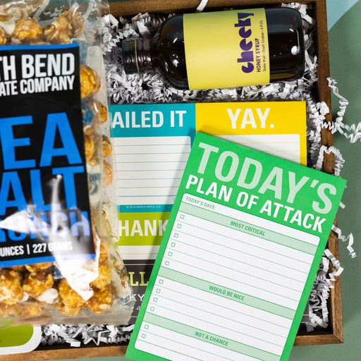 On a calming blue and green background with sunny yellow cutout sits a wooden tray of products and white paper krinkle. Each product has some sunny yellow, green or blue on the label. This photo is a close-up of "Today's Plan of Attack" check box note pad.