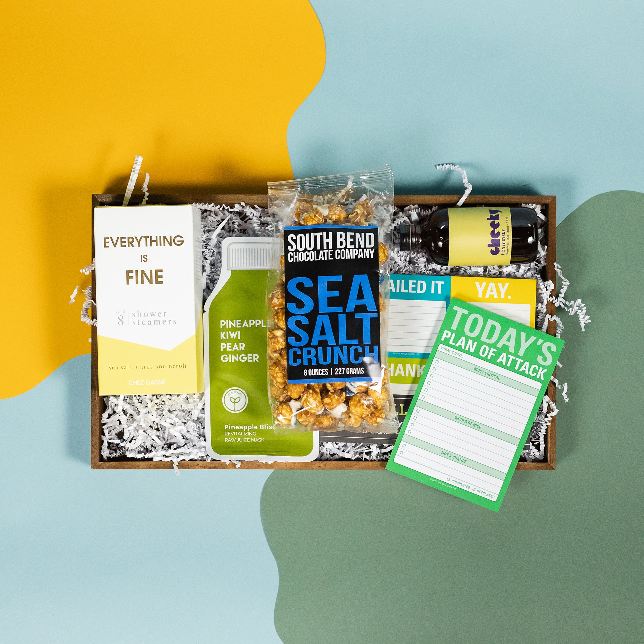 On a calming blue and green background with sunny yellow cutout sits a wooden tray of products and white paper krinkle. Each product has some sunny yellow, green or blue on the label. There's a pack of 8 Chez Gagne Sea Salt and Neroli "Everything is Fine" shower steamers, Pineapple Kiwi Pear Ginger Pineapple Bliss face mask, a mini bottle of Cheeky Honey Syrup, optimistic sticky note set, "Today's Plan of Attack" check box note pad, and a bag of South Bend Chocolate Company Sea Salt Crunch caramel corn.