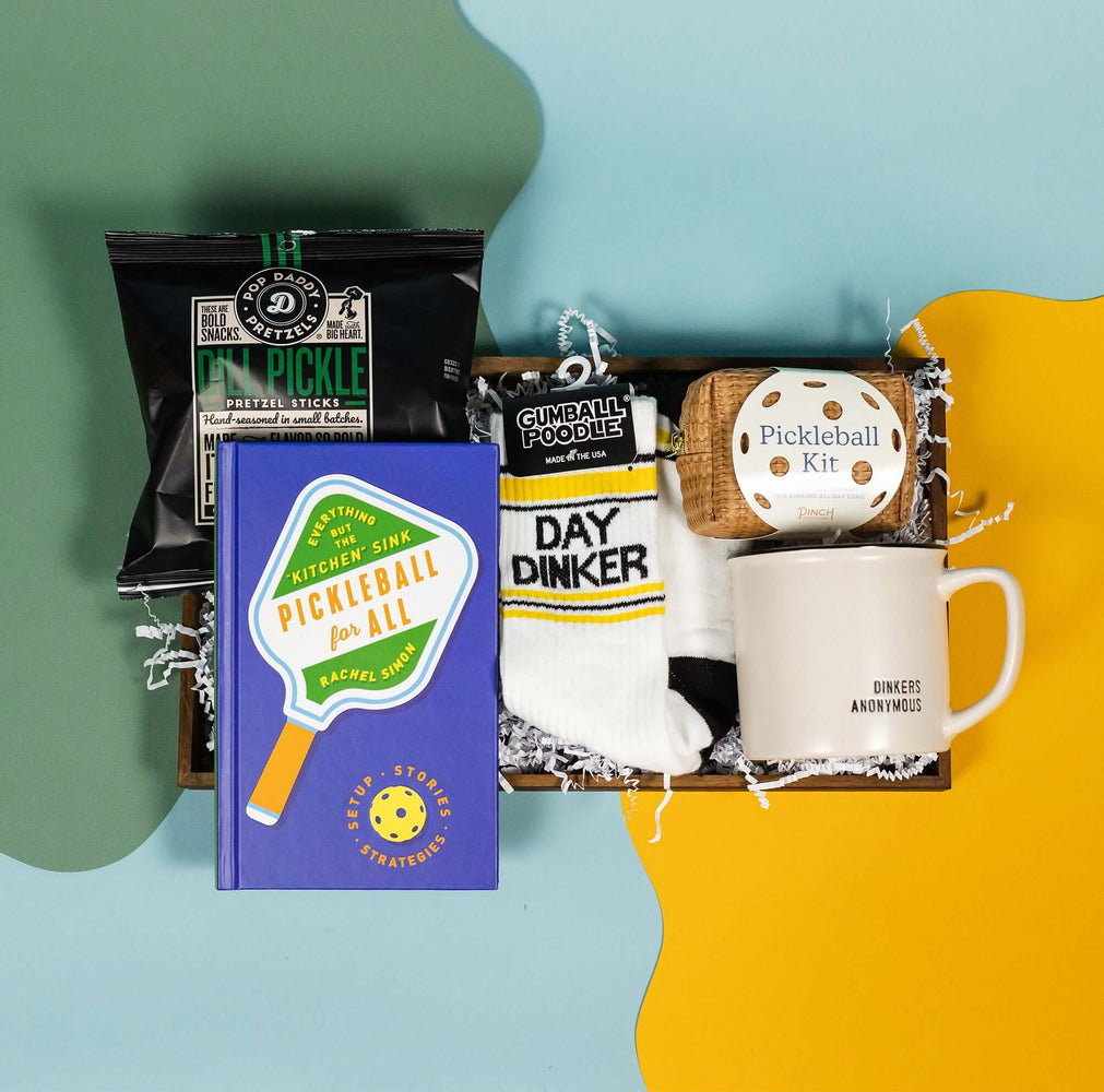 On a calming blue and green background with sunny yellow cutout sits a wooden tray of pickleball themed products and white paper krinkle. The products are the book "Pickleball for All: Setup, Stories & Strategies" book by Rachel Simon, Pop Daddy Dill Pickle Pretzel Sticks, Gumball Poodle "DAY DINKER" sporty novelty socks, a "Pickleball Kit" from Pinch Provisions, and an ivory mug with "DINKERS ANONYMOUS" embossed in black.