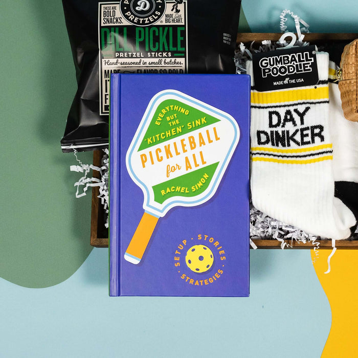 On a calming blue and green background with sunny yellow cutout sits a wooden tray of four pickleball themed products and white paper krinkle. This is a close-up of the book "Pickleball for All: Setup, Stories & Strategies" book by Rachel Simon, Pop Daddy Dill Pickle Pretzel Sticks, and Gumball Poodle "DAY DINKER" sporty novelty socks.
