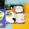 On a calming blue and green background with sunny yellow cutout sits a wooden tray of four pickleball themed products and white paper krinkle. This is a close-up of Gumball Poodle "DAY DINKER" sporty novelty socks, a "Pickleball Kit" from Pinch Provisions, and an ivory mug with "DINKERS ANONYMOUS" embossed in black.