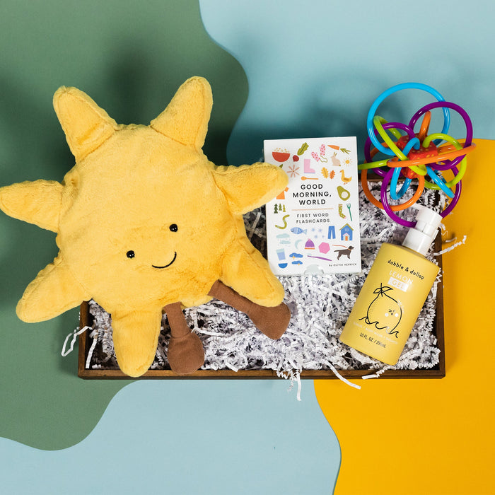 On a calming blue and green background with sunny yellow cutout sits a wooden tray of four baby themed products and white paper krinkle. The products are a yellow similing sun Jellycat plushie, a yellow bottle of Dabble & Dollop Lemon Gel tearless bubble bath, Good Morning, World First Word Flashcards, and Manhattan Toy Rainbow Rattle.