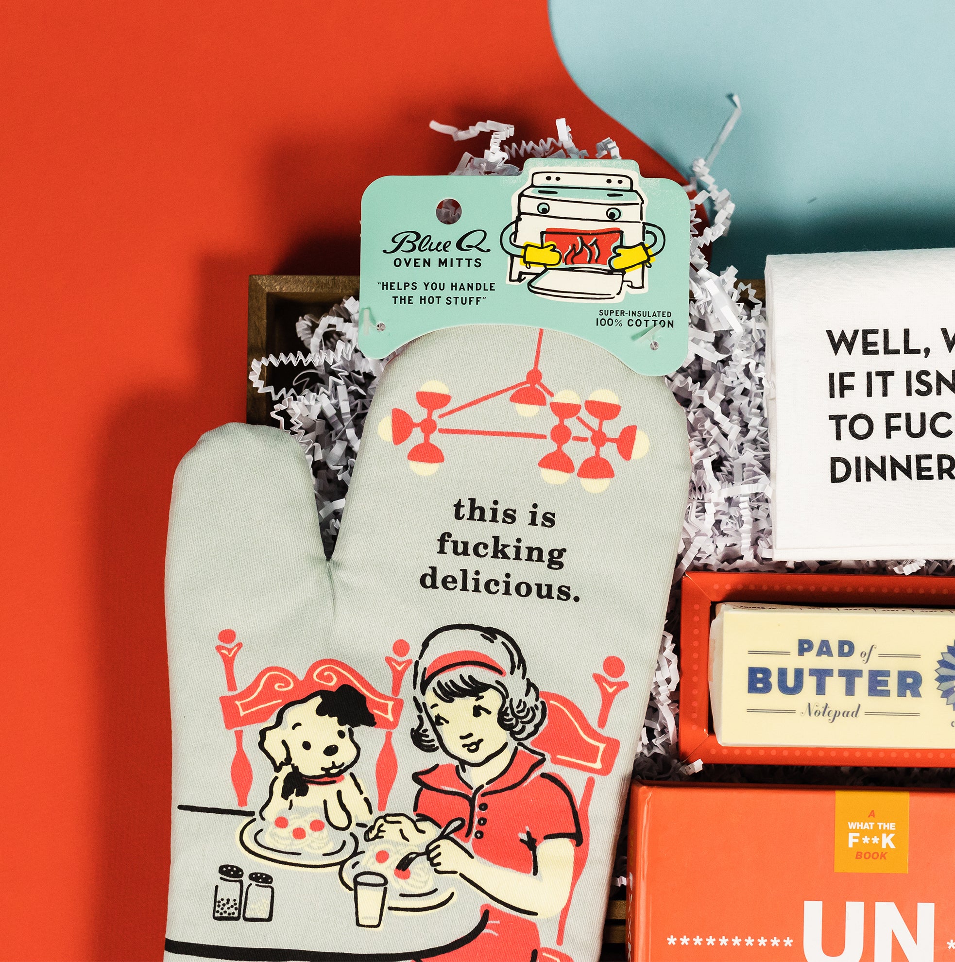 On a calming blue background with bold red wobbly cutout sits a wooden tray of snarky kitchen themed gifts among white paper krinkle. This photo is a close-up on the retro illustrated oven mitt that says "I'll Feed All You Fuckers," featuring a 50s style girl at the table eating with her little spotted puppy.