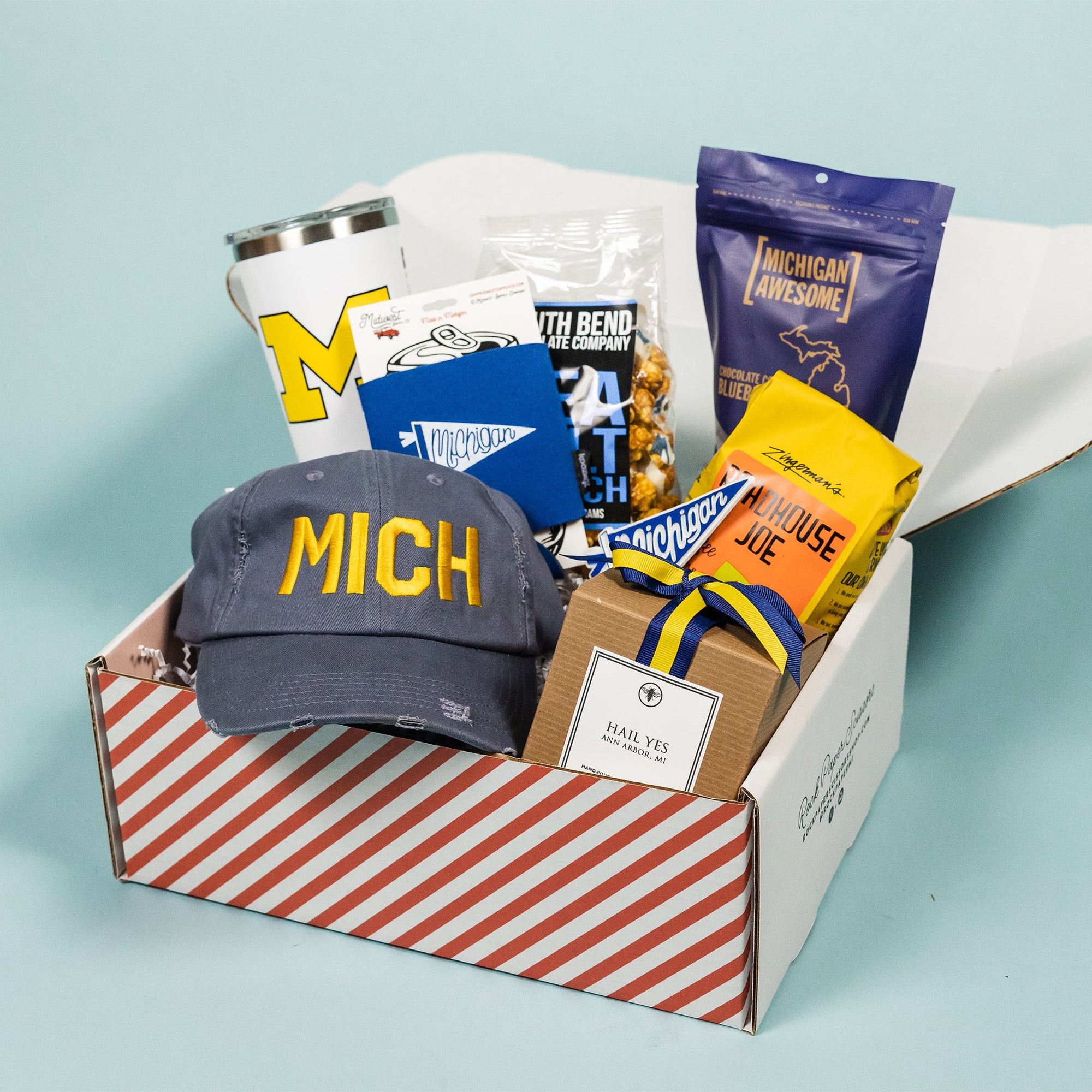 On a calming blue background sits a red and white striped box filled to the brim with U of M gifts. It includes a distressed navy ballcap that says "MICH" in maize, a 20 oz white liscensed block M Corkcicle, RPS exclusive "Hail Yes" hand-poured candle, Zingerman's Roadhouse Joe coffee beans, Michigan Awesome chocolate covered blueberries, South Bend Chocolate Sea Salt Caramel Corn, RPS Exclusive blue Michigan pennant koozie and blue pennant Michigan magnet.