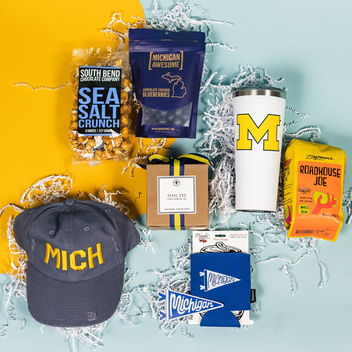 On a calming blue background with sunny yellow cutout lays an arrangement of Michigan gifts. Gifts include a distressed navy ballcap that says "MICH" in maize, a 20 oz white liscensed block M Corkcicle, RPS exclusive "Hail Yes" hand-poured candle, Zingerman's Roadhouse Joe coffee beans, Michigan Awesome chocolate covered blueberries, South Bend Chocolate Sea Salt Caramel Corn, RPS Exclusive blue Michigan pennant koozie and blue pennant Michigan magnet.