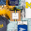 On a calming blue background with sunny yellow cutout lays an arrangement of Michigan gifts. This photo is a close-up of an RPS exclusive "Hail Yes" hand-poured candle, an RPS Exclusive blue Michigan pennant koozie, a distressed navy ballcap that says "MICH" in maize and a 20 oz white liscensed block M Corkcicle.
