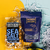 On a calming blue background with sunny yellow cutout lays an arrangement of Michigan gifts. This photo is a close-up of a navy bag of Michigan Awesome chocolate covered blueberries and clear bag of South Bend Chocolate Sea Salt Caramel Corn with a black and blue bold graphic label.