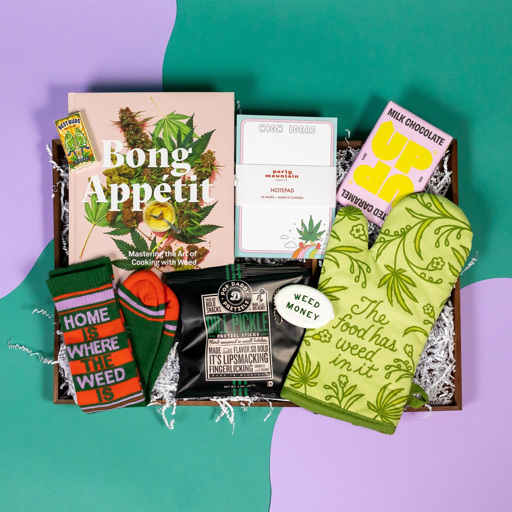 On a green and purple background sits a wooden tray of cannabis-themed gifts on white paper krinkle. The gifts include a "Bong Appetit" cookbook, "Home is Where the Weed Is" socks in kelly green, orange and lavender, a green illustrated oven mitt that says, "The Food Has Weed In It," "High Ideas" notepad with a cute cannabis cartoon, Pop Daddy Dill Pickle Popcorn, an UP UP Salted Caramel milk chocolate bar, a white coin keychain that says WEED MONEY in green and a tiny pack of novelty "Best Buds" gum.