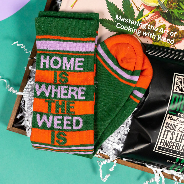 On a funky green and purple background sits a wooden tray of cannabis-themed gifts on white paper krinkle. This photo is a close-up on the "Home is Where the Weed Is" retro novelty socks. They're a funky dark green with orange and lavender stripes.