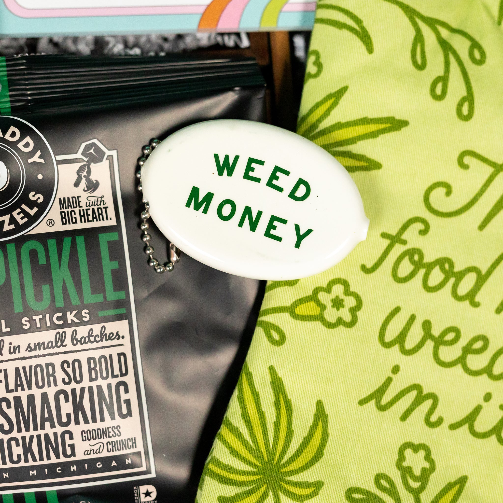 On a funky green and purple background sits a wooden tray of cannabis-themed gifts on white paper krinkle. This photo is a close-up of a small white coin keychain that says WEED MONEY in green.