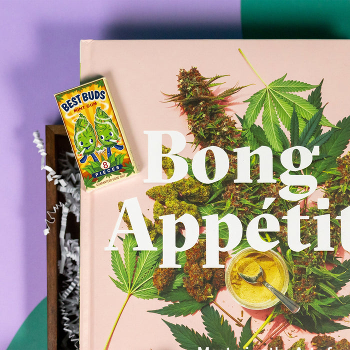 On a funky green and purple background sits a wooden tray of cannabis-themed gifts on white paper krinkle. This photo is a close-up on the Bong Appetit cookbook and tiny pack of "Best Buds" gum.
