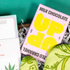 On a funky green and purple background sits a wooden tray of cannabis-themed gifts on white paper krinkle. This photo is a close-up of the UP UP Salted Caramel Milk Chocolate bar.