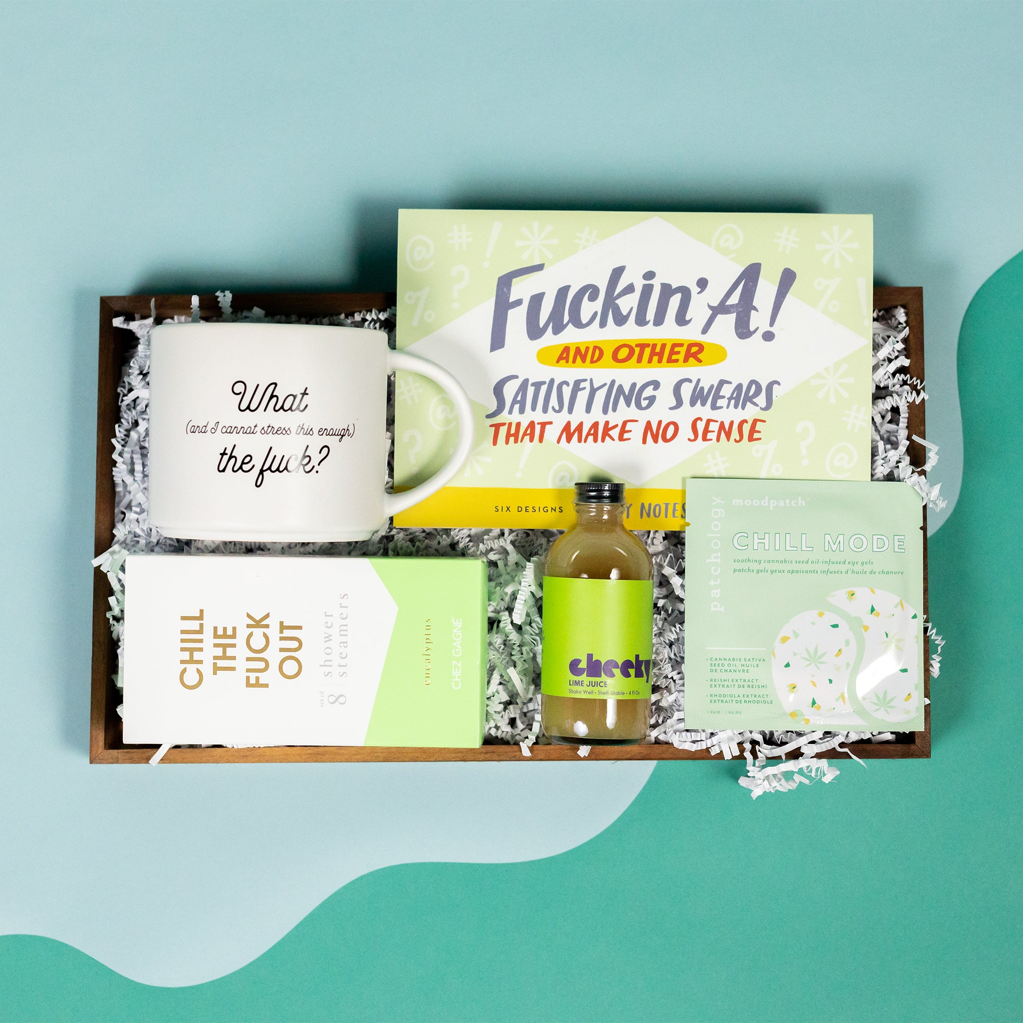 On a cool green and blue wavy background sits a wooden tray of gifts on white paper krinkle with light green themed products, including a white "What (and I cannot stress this enough) The Fuck" mug, "Fuckin' A! and other Satisfying Swears that Make No Sense" pack of sticky notes with 6 designs, an 8-pack of Chez Gagne eucalyptus "Chill the Fuck Out" Shower Steamers, a mini bottle of Cheeky Lime Juice for cocktails, and a Patchology Chill Mode soothing cannabis oil under eye gel patches.