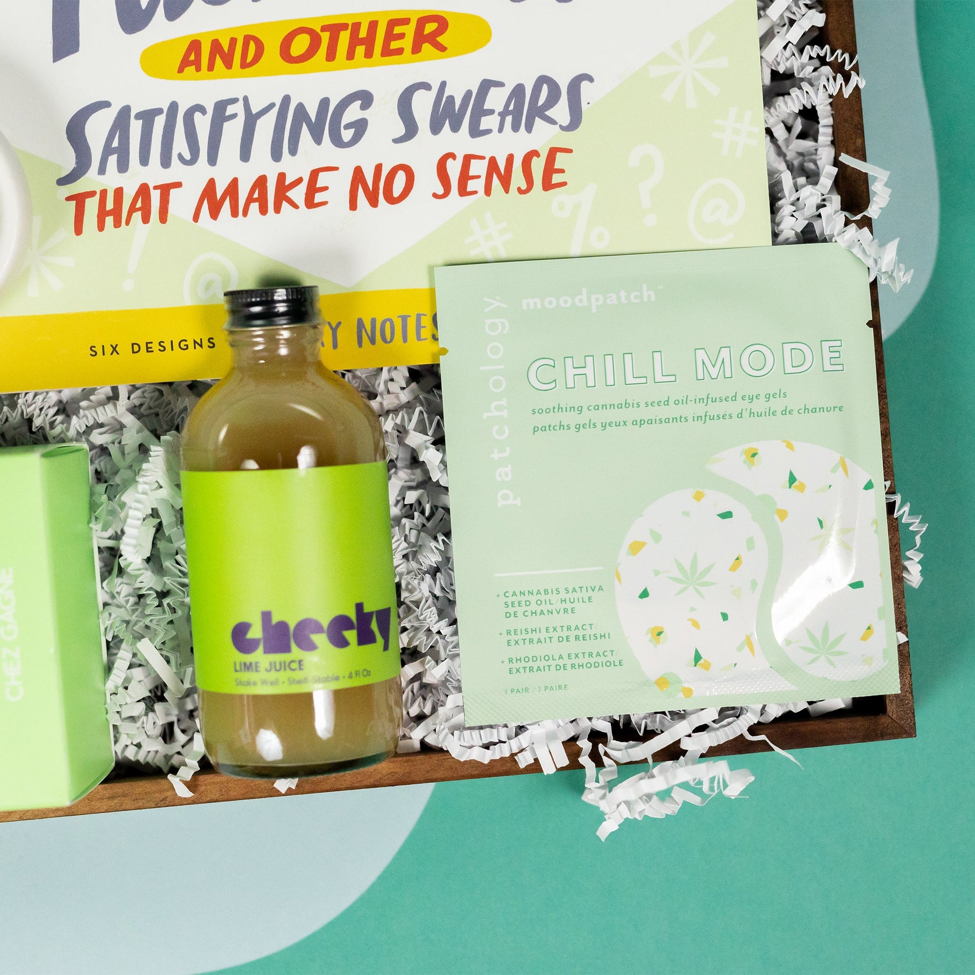 On a cool green and blue wavy background sits a wooden tray of gifts on white paper krinkle. This photo is a close-up on the mini bottle of Cheeky Lime Juice for cocktails, and a Patchology Chill Mode soothing cannabis oil under eye gel patches. Both have light green in the packaging and look cute together.