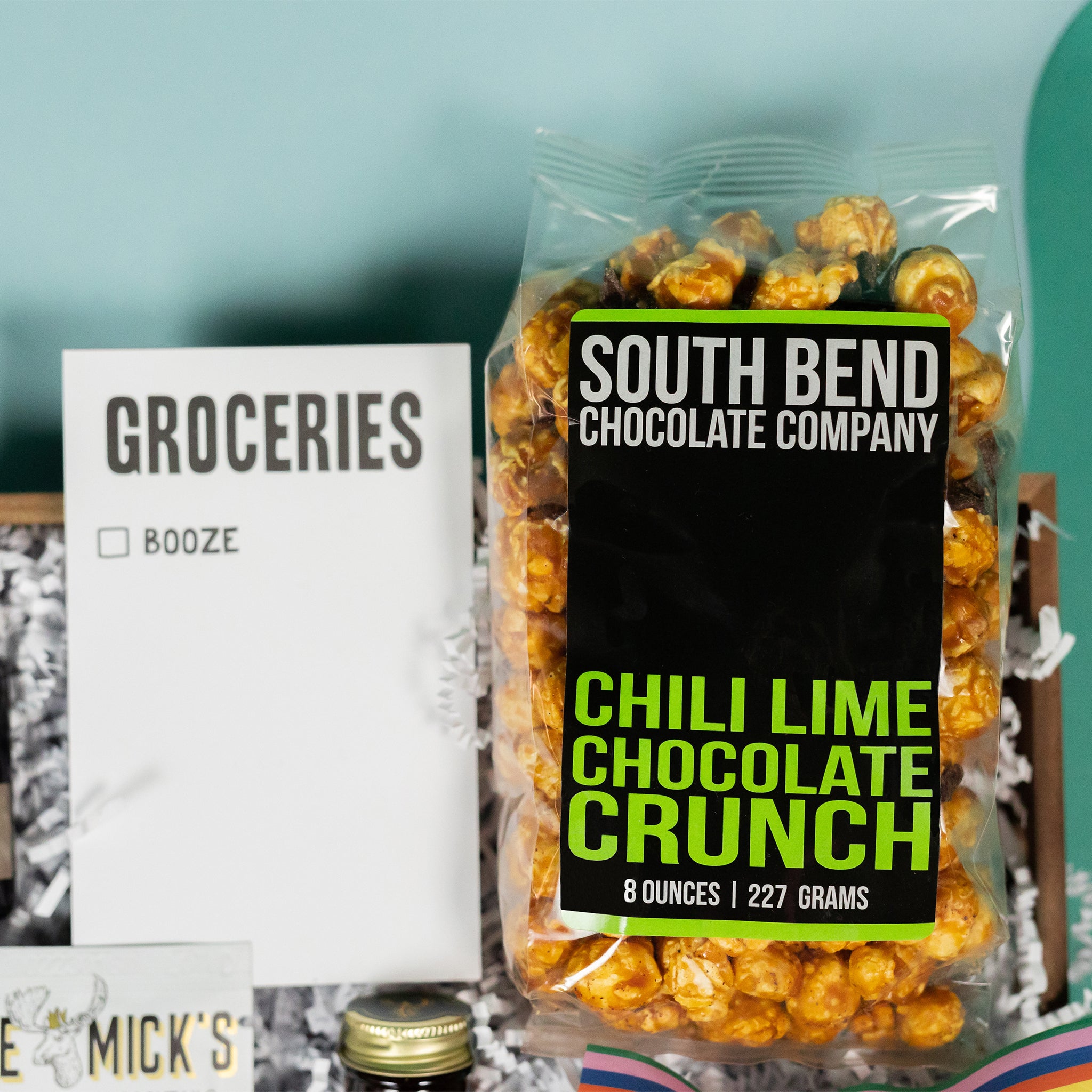 On a cool green and blue wavy background sits a wooden tray of moscow-mule themed gifts on white paper krinkle. This photo is a close-up on the  "Groceries" list with one checkbox for Booze and clear bag of South Bend Chocolate Company Chili Lime Chocolate Crunch Caramel Corn with a bold black and lime green graphic label.