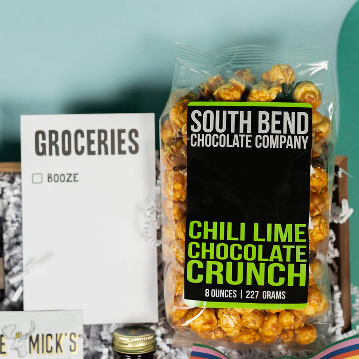 On a cool green and blue wavy background sits a wooden tray of moscow-mule themed gifts on white paper krinkle. This photo is a close-up on the  "Groceries" list with one checkbox for Booze and clear bag of South Bend Chocolate Company Chili Lime Chocolate Crunch Caramel Corn with a bold black and lime green graphic label.