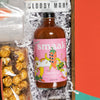 On a funky green background with raspberry and tomato red wobbly cutout shapes sits a wooden tray of bloody-mary themed gifts on bright white krinkle paper. Just add vodka! This photo is a close-up on the Smaak Cayenne hot sauce. The label has a trippy pink illustration of a hot pepper gettin' down with a boombox and his bad self.