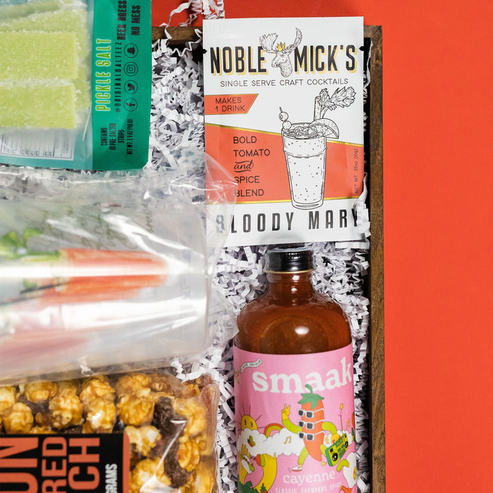 On a funky green background with raspberry and tomato red wobbly cutout shapes sits a wooden tray of bloody-mary themed gifts on bright white krinkle paper. Just add vodka! This photo is a close-up on the Noble Mick's Single Serve Bloody Mary Craft Cocktail.
