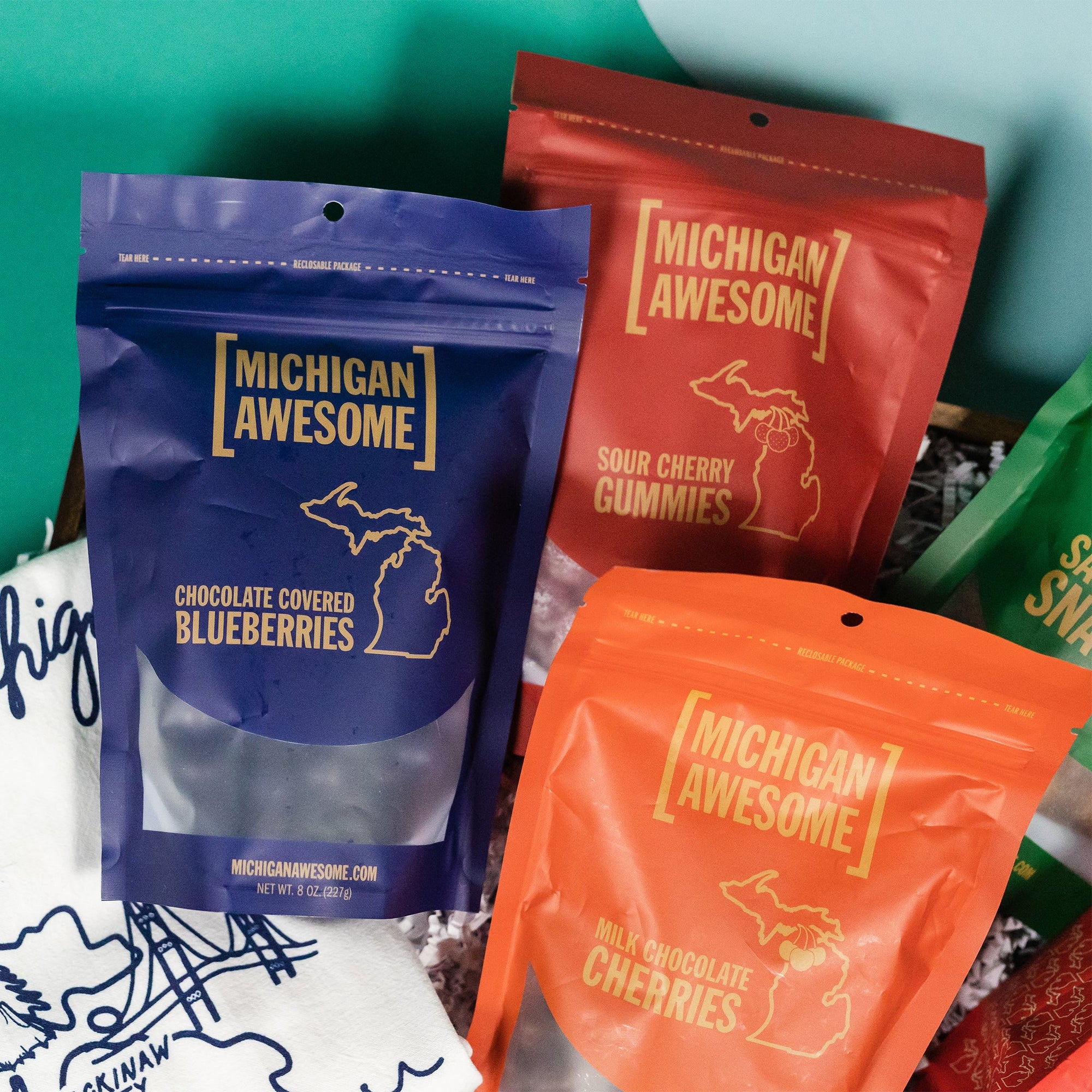 On a calming blue and teal background sits a wooden tray with an assortment of Michigan Awesome snacks and an illustrated map of Michigan tea towel. This photo is a close-up of Michigan Awesome Chocolate Covered Blueberries, Sour Cherry Gummies, and Milk Chocolate Cherries.