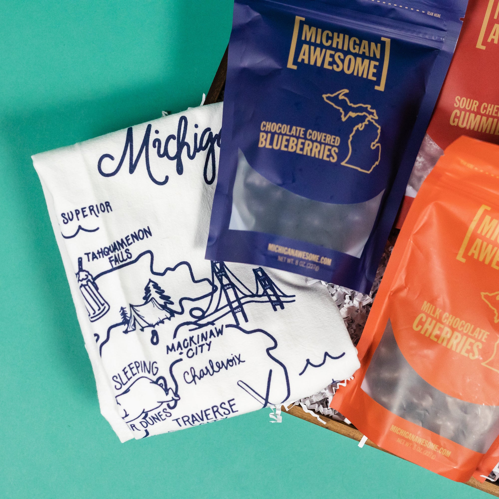 On a calming blue and teal background sits a wooden tray with a close-up of an assortment of Michigan Awesome snacks and an illustrated map of Michigan tea towel.