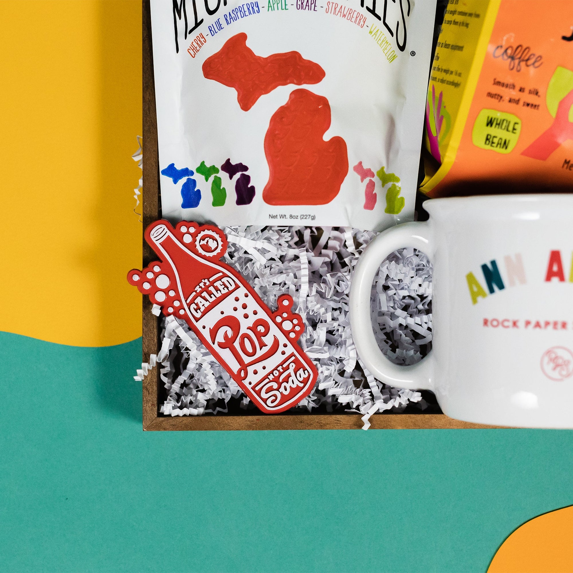 On a bright kelly green background with sunny yellow and orange wobbly cutouts sits a wooden tray with an assortment of Michigan and Ann Arbor themed gifts. This photo is a close-up of the "It's Called Pop Not Soda" red fridge magnet.
